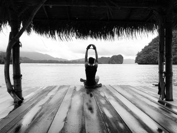 Man with arms raised sitting on wood by sea against sky