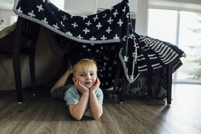 Portrait of confident boy with hands on chin lying under blanket tent at home