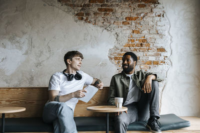 Young entrepreneur discussing with male colleague while sitting on bench against brick wall