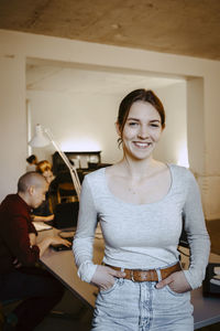 Portrait of smiling businesswoman with hands in pockets while colleague working at desk in office