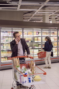Mature man talking on mobile phone while woman looking at display cabinet in refrigerated section of supermarket
