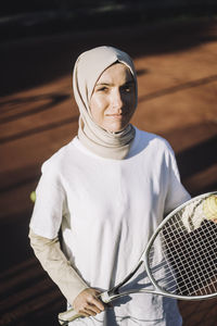 Confident woman wearing headscarf holding tennis racket on sunny day