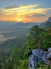 Man sitting on cliff at forest during sunset