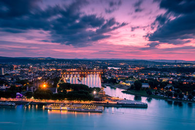 Illuminated buildings by river against sky at sunset