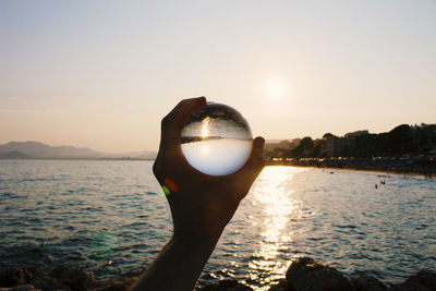 Cropped hand holding crystal ball against sea and sky during sunset