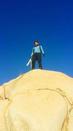 Low angle view of man holding knife while standing on rock against clear blue sky