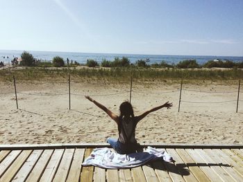 Rear view of woman sitting on floorboard looking at beach against sky