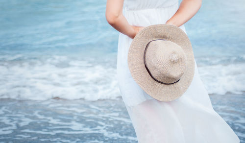 A young girl in a white dress stands with her back  of the blue sea, holding a light hat behind her. 