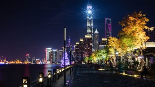 Illuminated modern buildings by river against sky at night