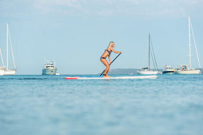 Side view of fit female surfer in bikini standing on paddleboard and rowing while training in sea on sunny day