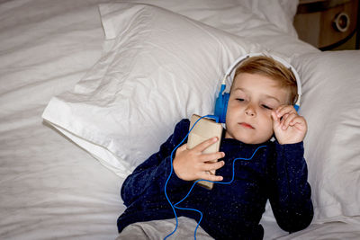 Small kid using mobile phone while wearing headphones and resting on the bed.