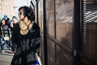 Young woman in warm clothing wearing sunglasses standing by built structure