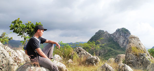 Side view of young man sitting on rock against sky