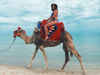 Portrait of young woman riding camel at beach against cloudy sky