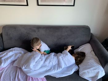 High angle view of siblings using wireless technology while lying on sofa at home