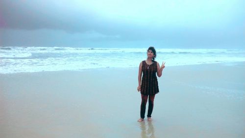 Portrait of young woman showing peace sign while standing at beach against cloudy sky