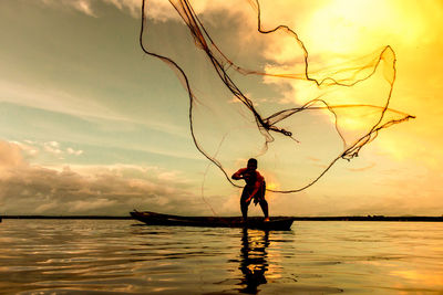 Man throwing fishing net in lake against sky during sunset