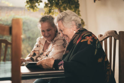 Elderly sisters inspecting pictures in photo album and discussing memories while sitting at table at home together