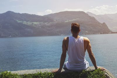 Rear view of man sitting while looking at sea and mountains