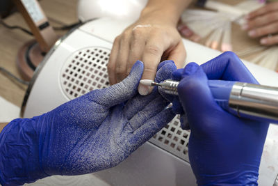 The manicurist grinds the nail plate with a milling machine in blue latex gloves.