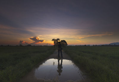 Man reflecting in puddle while standing amidst grassy landscape against sky during sunset