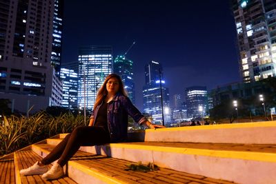 Full length of woman sitting against illuminated buildings in city at night