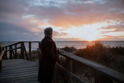 Side view of anonymous elderly female traveler in casual clothes standing on wooden pier on sandy beach and enjoying seascape at sunset