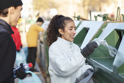 Smiling female environmentalist with friend throwing waste in garbage can