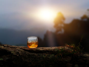 Close-up of glass on rock against sky during sunset