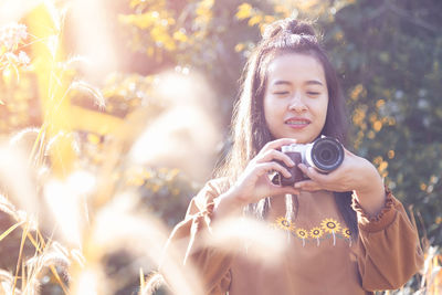 Smiling woman holding camera while standing against tree