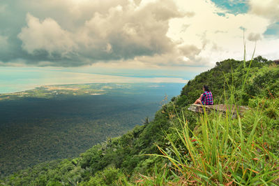 Side view of young woman sitting on mountain against cloudy sky