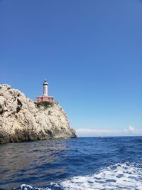 Lighthouse on rock by sea against clear blue sky