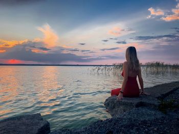 Rear view of woman in red dress sitting down near the lake at sunset and looking at the sky