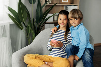 Sister and younger brother are sitting at home in an armchair and playing on the phone.