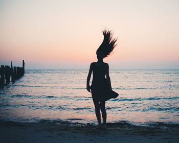Silhouette woman tossing hair while standing at beach against sky during sunset