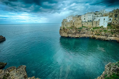 Buildings in rock by sea against cloudy sky