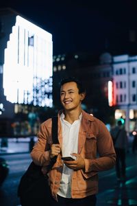 Smiling young man looking away with phone while standing in illuminated city at night