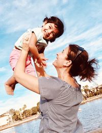 Smiling mother carrying daughter while standing at lake against sky