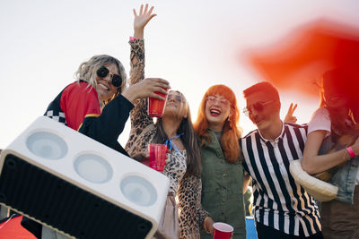 Happy friends with speaker and drinks enjoying in music festival against sky