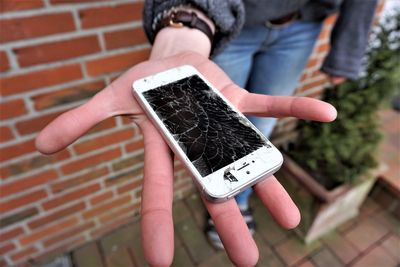 Low section of person holding damaged smart phone against brick wall