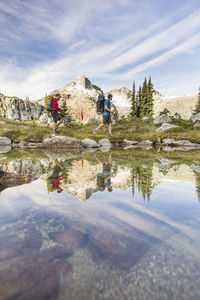 Side view and reflection of backpackers hiking beside alpine lake.