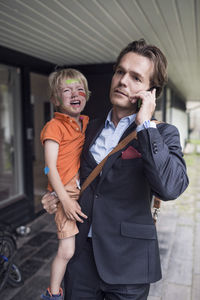 Businessman using mobile phone while carrying crying son outside house