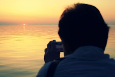 Rear view of man photographing sea against sky during sunset