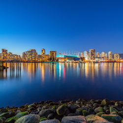 Illuminated buildings by river against blue sky