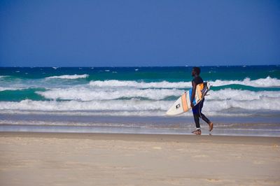 Full length of a man walking at beach against clear sky with his surfing board 