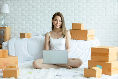 Full length of young woman using laptop while sitting by bed at home