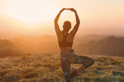 Rear view of woman meditating on field against sky during sunset