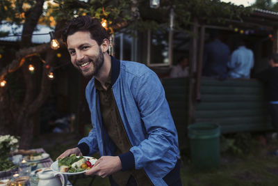 Portrait of happy man holding food plate while standing by dinner table at yard