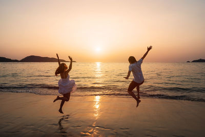 Carefree couple with arms raised jumping at beach against clear sky during sunset