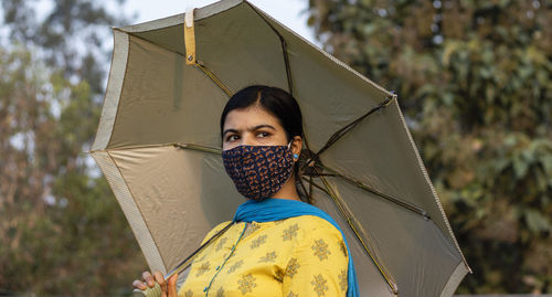 An indian woman in yellow dress and face mask on is going outside with umbrella during corona virus